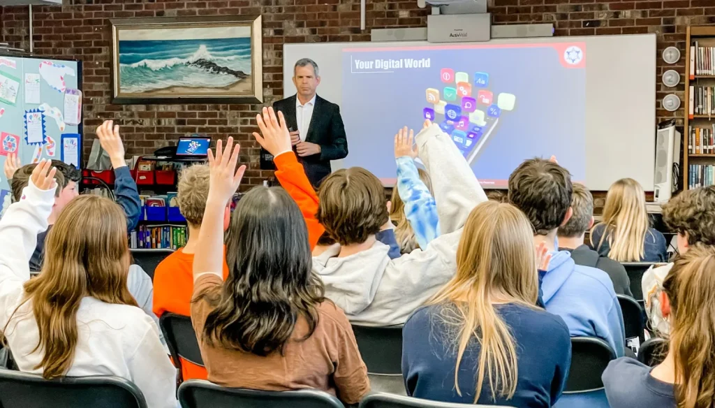 A man in a suit stands in front of a classroom presenting a slide titled "Your Digital World" as several students raise their hands, eager to learn about Digital Safety from the Cyber Safety Cop.