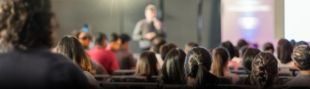 A blurred speaker stands at the front of a room addressing an audience seated in rows, suggesting a seminar for parents focused on online safety. The focus is on the backs of the attentive audience members' heads.