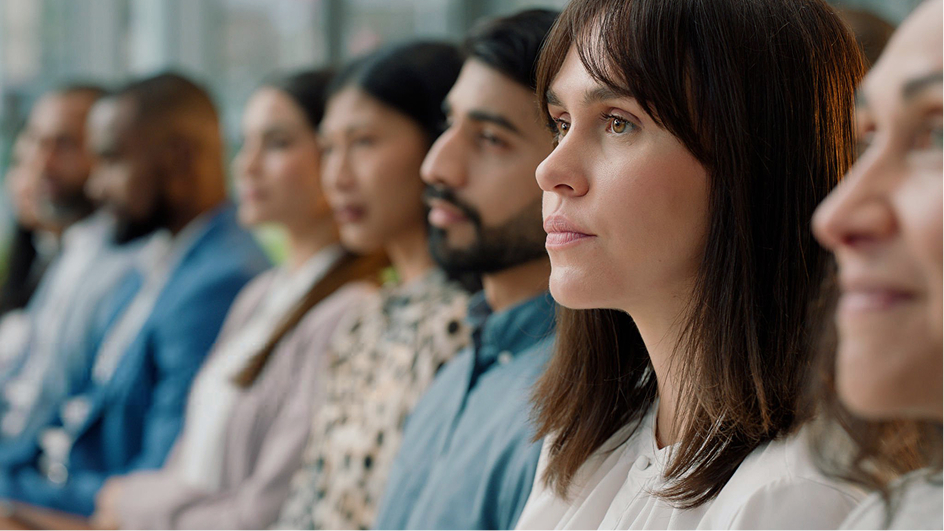 A diverse group of people sits in a row, attentively listening to keynote engagements off-camera. The focus is on a woman in the foreground with brown hair and a serious expression, while the background remains softly blurred.