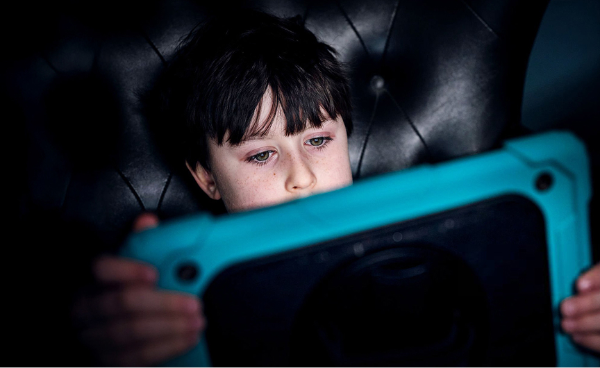A young boy with dark hair sits on a dark, tufted chair, intently looking at a blue tablet he is holding in front of him—his focused expression reminiscent of the concentration seen during keynote engagements. The dim background highlights his focus.