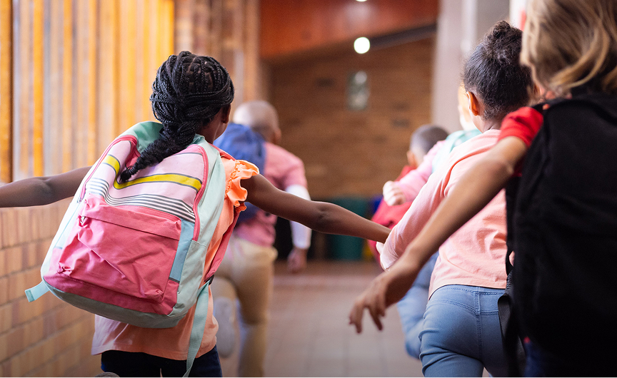 Children wearing backpacks run excitedly down a school hallway with their backs to the camera, as sunlight streams through large windows on the left, brightening the brick wall—capturing a moment as lively as Keynote Engagements.