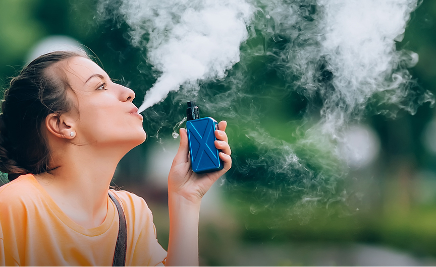 A person outdoors holds a blue vape device, blowing a large cloud of vapor. The blurred green background suggests a park or natural setting—perhaps a break during Keynote Engagements. The individual wears a yellow shirt and has hair tied back.