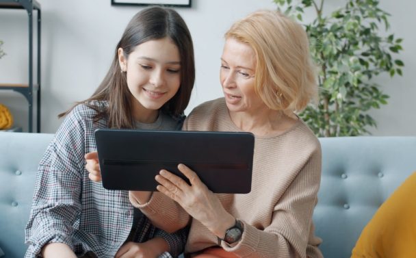 Two women sit on a couch, smiling as they explore AI Chatbots on a tablet together. The younger woman has long dark hair, the older one has short blonde hair. Green plants and shelves decorate the background.